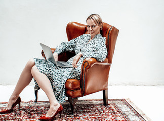 Relaxed young adult female model sitting in a leather chair with a laptop on her knees, looking casual