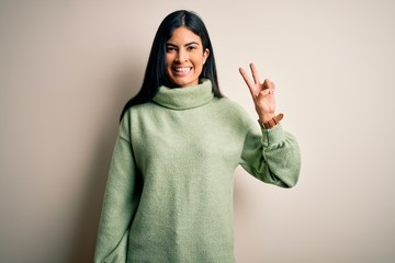 Young beautiful hispanic woman wearing green winter sweater over isolated background showing and pointing up with fingers number two while smiling confident and happy.