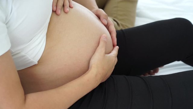 the young asian pregnancy woman with  husband sitting on the white bed with the hands touching the bally with love and care 