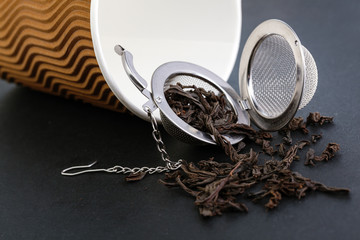 Black tea and a tea strainer with a chain, on the background of an eco-friendly paper cup, lie on a black table. A tea strainer is a kitchen accessory,  closeup.