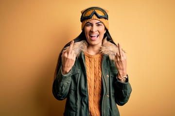 Young beautiful hispanic woman wearing ski glasses and coat for winter weather Showing middle finger doing fuck you bad expression, provocation and rude attitude. Screaming excited