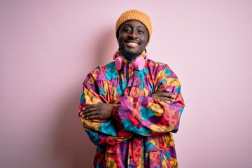 Young handsome african american man wearing colorful coat and cap over pink background happy face smiling with crossed arms looking at the camera. Positive person.