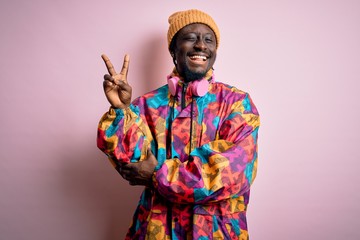 Young handsome african american man wearing colorful coat and cap over pink background smiling with...