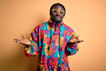 Handsome african american man wearing colorful coat and glasses over yellow background clueless and confused expression with arms and hands raised. Doubt concept.