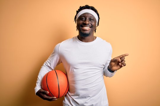 Young african american sportsman doing sport holding basketball ball over yellow background very happy pointing with hand and finger to the side