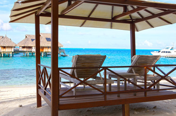 chairs on the sandy beach and houses over water of the blue sea. Polynesia.