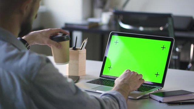 man working on laptop with green screen at office. Spbd he drinks coffee from paper cup. chroma key computer for business chroma key purposes. concept break, hands typing