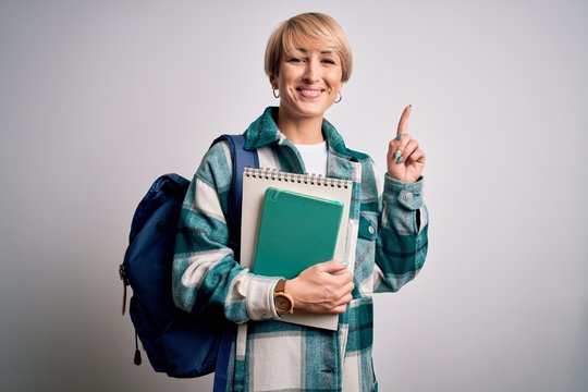 Young blonde student woman with short hair wearing backpack and holding university books surprised with an idea or question pointing finger with happy face, number one