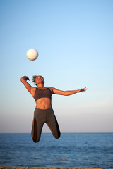Stunning young woman serving volleyball at sunset on a beach with ocean behind her