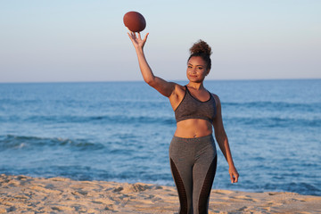 Stunning young woman throwing an American football on beach with ocean behind her