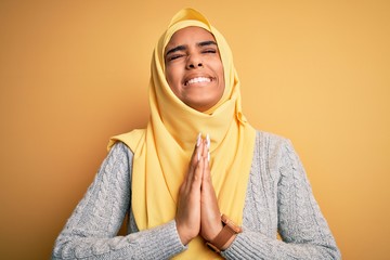 Young beautiful african american girl wearing muslim hijab over isolated yellow background begging and praying with hands together with hope expression on face very emotional and worried. Begging.