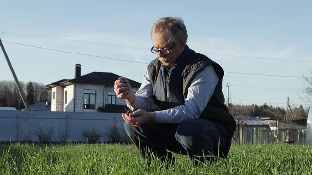 An elderly man in a blue vest and glasses squats. With his hands he squeezes, wrinkles and pours the earth. He is in his garden. Assesses soil readiness. Warm evening light. The average plan. UHD.