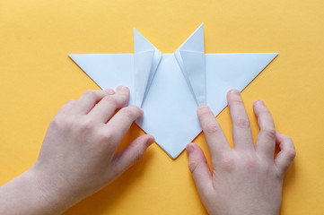 Step 12. Hands of a child on a yellow background divide a square of white paper into triangles. Origami bull concept.