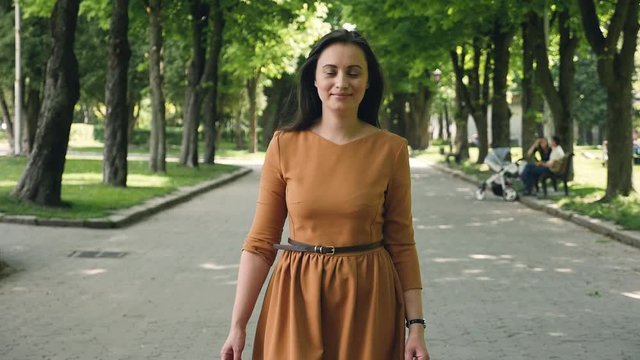 Portrait of a Young Beautiful Brunette Woman walking in the park. Girl is Dressed in Orange dress and is Walking on the Edge of the Forest. She Smiles and looking to camera.