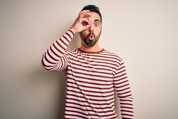 Young handsome man with beard wearing casual striped t-shirt standing over white background doing ok gesture shocked with surprised face, eye looking through fingers. Unbelieving expression.