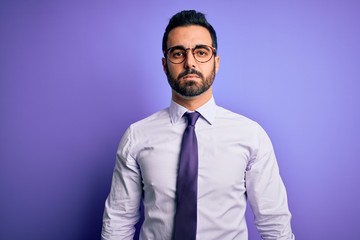 Handsome businessman with beard wearing casual tie and glasses over purple background with serious expression on face. Simple and natural looking at the camera.