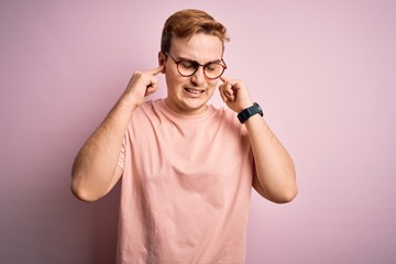 Young handsome redhead man wearing casual t-shirt standing over isolated pink background covering ears with fingers with annoyed expression for the noise of loud music. Deaf concept.