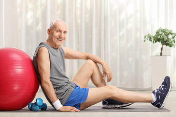 Elderly man with fitness ball and dumbbells sitting on an exercise mat