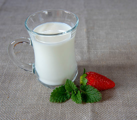 A Cup of milk , a strawberry berry, and mint leaves on a gray linen tablecloth.