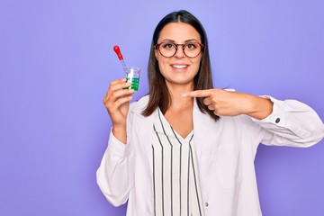 Young beautiful brunette scientist woman wearing coat and glasses holding test tube Smiling happy pointing with hand and finger
