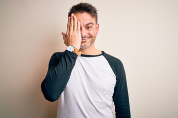 Young handsome man wearing casual t-shirt standing over isolated white background covering one eye with hand, confident smile on face and surprise emotion.