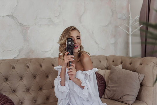Beautiful girl in a white dress having fun with an old camera on the sofa. The bride poses and laughs at home. Light and professional makeup and airy curls with fashion accessories