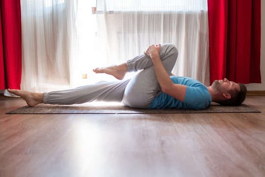 Caucasian man doing yoga on the floor, practicing the Supported One Legged Wind Relieving Pose or Eka Pada Pavanamuktasana at home in the living room.