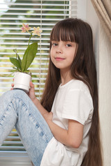 A child is sitting on a windowsill with a flower in his hands. A little girl (seven years old) is sitting on a windowsill by the window and holding a pot of orchid flower. One person