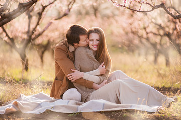 A young man in a brown light jacket gently embraces a beautiful girl with long hair in the back - a couple sitting in nature in a flowering garden. selective front and back focus