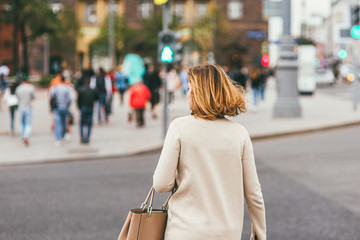 A young woman with white hair, wearing a light beige coat and carrying a leather purse, crosses a pedestrian crossing. Selective focus macro shot with shallow DOF. Social distancing concept photo