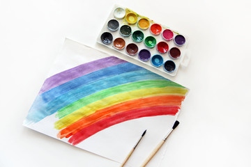 A brush, a set of paints, and a sheet of paper with a painted rainbow on a white table.
