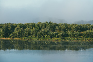 Early morning l Spring landscape with a full flowing river in the foreground and a green Bank overgrown with bushes and trees in the background. The distant plan is drowned in a thick morning fog