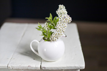 A branch of blooming spirea in a white milkman on a vintage stool opposite a dark blue wall