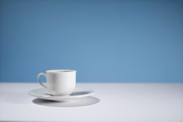 White cup of morning espresso on a stone surface of a table with reflections opposite blue background.