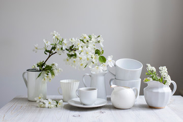 Vase with a blossoming branch of an apple tree. A set of white dishes: a cup of coffee, milk jug, a jug, a tray on the table against the background of a blue wall.