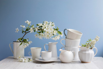Vase with a blossoming branch of an apple tree. A set of white dishes: a cup of coffee, milk jug, a jug, a tray on the table against the background of a blue wall.