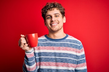 Young blond man with curly hair drinking cup of coffee standing over red background with a happy face standing and smiling with a confident smile showing teeth