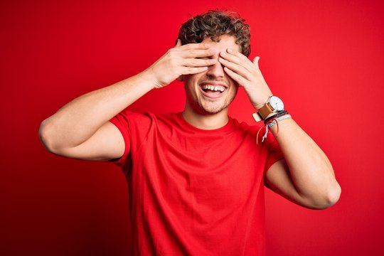 Young blond handsome man with curly hair wearing casual t-shirt over red background covering eyes with hands smiling cheerful and funny. Blind concept.