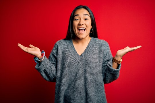 Young beautiful asian woman wearing casual sweater standing over isolated red background celebrating crazy and amazed for success with arms raised and open eyes screaming excited. Winner concept