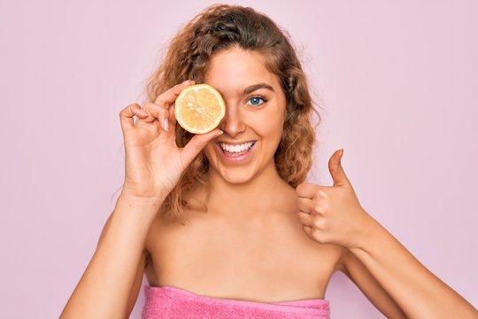 Beautiful woman with blue eyes wearing towel shower after bath holding lemon fruit over eye happy with big smile doing ok sign, thumb up with fingers, excellent sign
