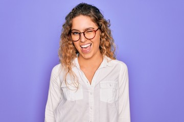 Young beautiful woman with blue eyes wearing casual shirt and glasses over purple background winking looking at the camera with sexy expression, cheerful and happy face.