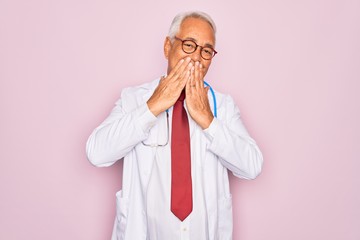 Middle age senior grey-haired doctor man wearing stethoscope and professional medical coat laughing and embarrassed giggle covering mouth with hands, gossip and scandal concept