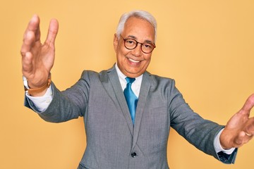 Middle age senior grey-haired handsome business man wearing glasses over yellow background looking at the camera smiling with open arms for hug. Cheerful expression embracing happiness.