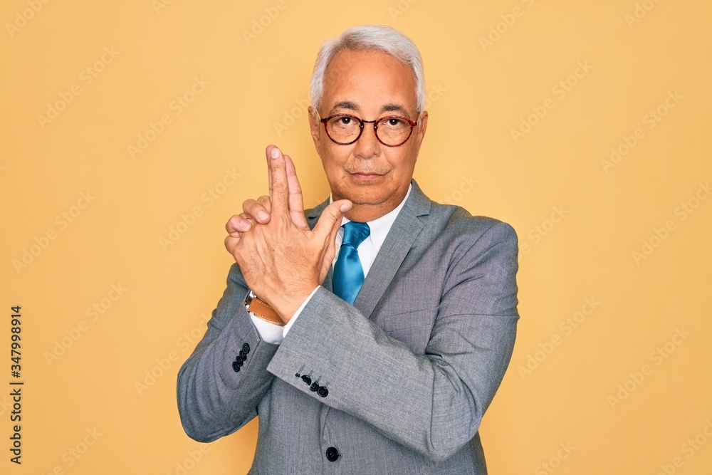 Sticker Middle age senior grey-haired handsome business man wearing glasses over yellow background Holding symbolic gun with hand gesture, playing killing shooting weapons, angry face