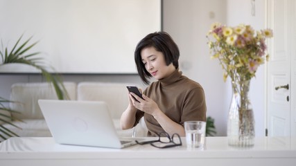 Asian woman in eyeglasses working with laptop in white home office and browsing internet in smartphone.