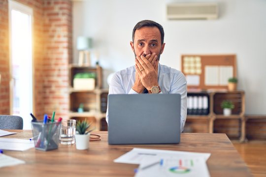 Middle age handsome businessman wearing tie sitting using laptop at the office shocked covering mouth with hands for mistake. Secret concept.