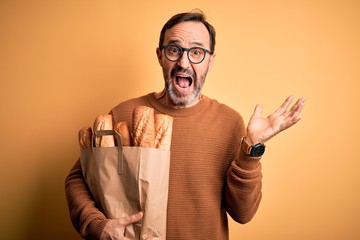 Middle age hoary man holding paper bag with bread standing over isolated yellow background very happy and excited, winner expression celebrating victory screaming with big smile and raised hands