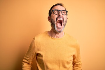 Middle age hoary man wearing casual sweater and glasses over isolated yellow background angry and mad screaming frustrated and furious, shouting with anger. Rage and aggressive concept.