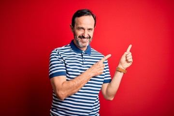 Naklejka premium Middle age hoary man wearing casual striped polo standing over isolated red background smiling and looking at the camera pointing with two hands and fingers to the side.
