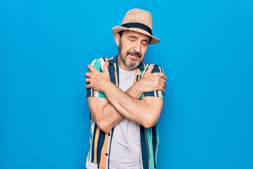 Middle age handsome tourist man on vacation wearing shirt and hat over blue background hugging oneself happy and positive, smiling confident. Self love and self care